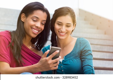 Closeup shot of young woman texting on mobilephone with her friend near. Happy smiling female friends looking at smart phone. African woman showing the smartphone to her friend outdoor.