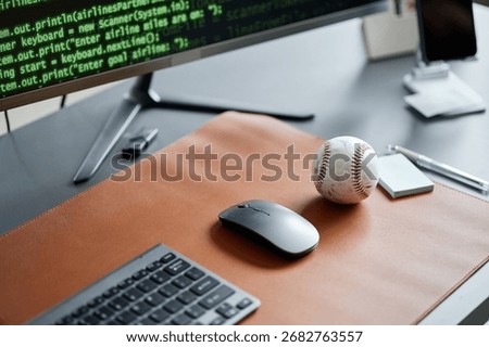 Closeup showing computer keyboard, wireless mouse, baseball, and monitor displaying programming code on desk, illustrating workplace of software developer with sports interest