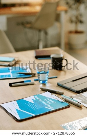 Closeup showing documents, pens, digital tablet, glass of water on wooden table in modern office, suggesting business meeting or real estate planning, no people visible
