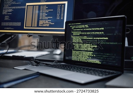 Closeup showing laptop with green code on screen positioned in front of desktop monitor displaying command line interface, illustrating software development or cybersecurity workspace