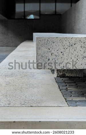 A close-up shows empty, modern stone benches with a textured finish. The benches vary in height and sit on a paved surface, suggesting a public or urban space. Clean lines and neutral tones.