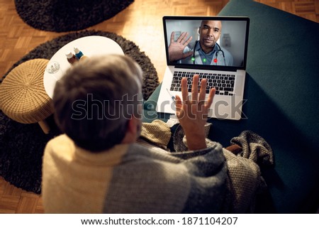 Close-up of sick man waving while calling his doctor via video call at night from home. Focus is on doctor on laptop screen.
