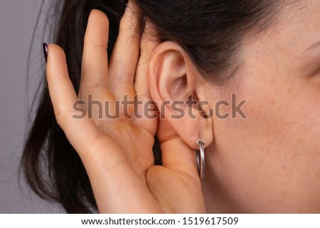 A closeup and side profile view of a young caucasian woman struggling to hear the conversation, gesturing with her hand behind her ear to speak up, wax build up and deafness.