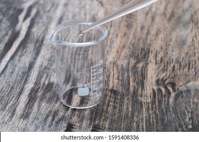 Close-up side view of 25 ml chemistry empty measuring beaker with a pipette on the wooden surface, a drop of transparent liquid in a plastic transparent slender tube - Powered by Shutterstock