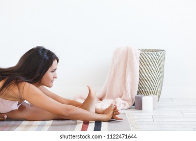 Close-up Side View Of Charming Cute Young Brunette Woman A Yoga Instructor Doing An Exercise Maha Mudra Sitting On Rug In A White Room. Place For Text