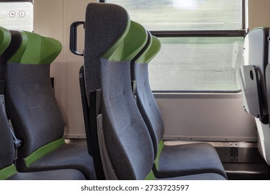 A close-up side view of empty gray and green fabric passenger seats inside a clean, modern commuter train or tram next to a large window during the day.