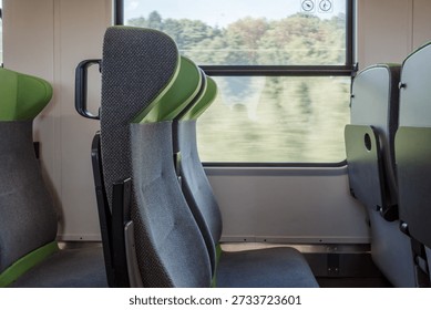 A close-up side view of empty gray and green fabric passenger seats inside a clean, modern commuter train or tram next to a large window during the day.