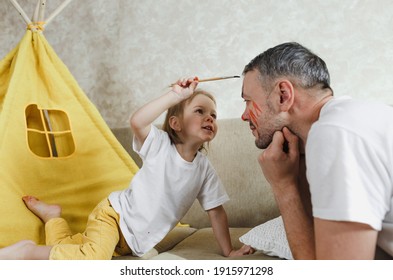 Close-up Side View Of A Happy Little Girl Applying Paint Brush To Her Father's Face. A Handsome Smiling Male Father Plays With His Daughter On The Couch At Home.