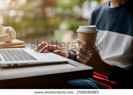 A close-up side view image of a man working remotely from an outdoor table, using his laptop computer while holding a coffee cup. people and technology concepts