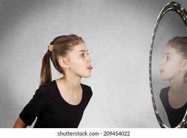 Closeup Side View Profile Portrait Child, Girl Sticking Her Tongue Out Looking In The Mirror, Isolated Grey Wall Background. Human Facial Expressions, Emotions, Feelings, Life Perception Body Language