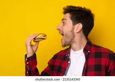 Closeup Side View Profile Portrait Of Funny Hungry Guy Biting Burger Eating Junk Food Holding Sandwich With Open Mouth Posing On Yellow Studio Background. Man Enjoying Big Hamburger, Bad Eating Habit