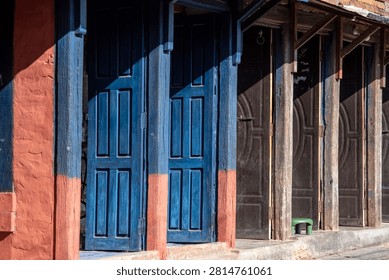 A close-up, side-angle view showing a row of traditional wooden doors along a village street facade in Nepal. On the left, vibrant blue paneled doors stand out against orange-terracotta painted pillars, while the rest of the row features dark, carved wooden doors framed by weathered rustic pillars.