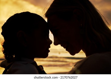 Closeup silhouette of a mother and daughter standing face to face on the beach at sunset. Backlit young woman and her girl child smiling and facing one another with the ocean in the background