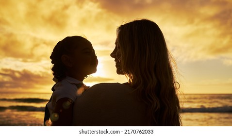 Closeup silhouette of mother and daughter standing on the beach at sunset. Backlit young woman and girl child smiling and talking with the ocean in the background. Single parent and kid on vacation