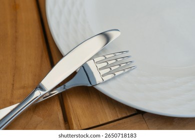 a close-up of a silver brand knife and fork resting on the edge of a white ceramic plate with a subtle textured pattern, set on a warm wooden table. this image is suitable for use in cutlery, dining. - Powered by Shutterstock - Get 15% off with code: PIKWIZARD15