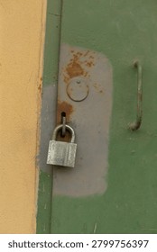 Close-up of a silver padlock securing a worn green metal door next to a yellow painted wall. The door shows patches of rust, scratched paint and a recessed