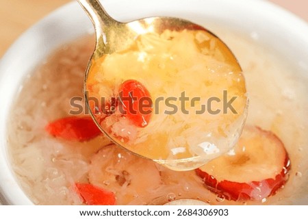 Close-up of silver spoon lifting clear broth soup with red goji berries and mushroom slices from white bowl, showing traditional Asian cuisine preparation