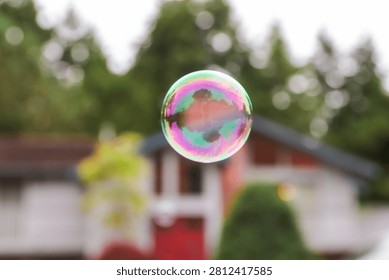 Close-up of a single iridescent soap bubble floating outdoors with a blurred suburban house and trees in the background, featuring colorful reflections, soft bokeh, and shallow depth of field.