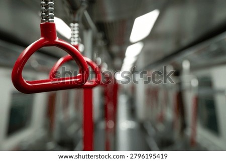 A close-up of a single red triangular hanging handle suspended from a braided chrome fitting inside an MTR train carriage in Hong Kong. Further handles and the vertical red pole recede along the length of the carriage in the blurred background, alongside fluorescent ceiling panels and white interior walls. The MTR is Hong Kong's mass transit railway network, serving over five million passenger journeys daily.