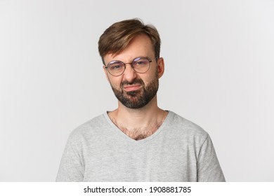 Close-up of skeptical bearded man in glasses and gray t-shirt, grimacing disappointed, standing over white background
