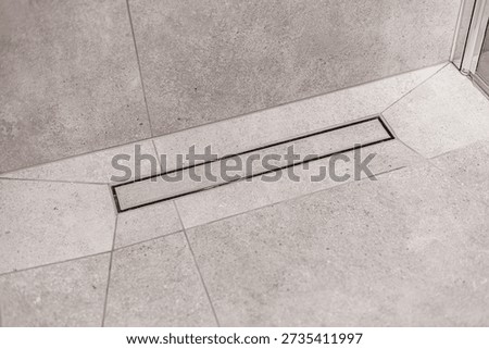 Close-up of a sleek linear drain integrated into a light gray stone-tiled shower floor. Clean, geometric lines emphasize modern bathroom design
