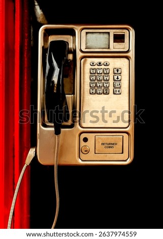A close-up, slightly high-angle view of a public payphone attached to a vibrant red structure. The payphone, metallic gold in color, features a keypad, a small display screen, and a coin return slot.