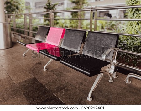 ​A close-up, slightly low-angle view of four empty metal waiting chairs, featuring one bright pink seat among three black ones, set on a dark tiled floor outside a modern building.