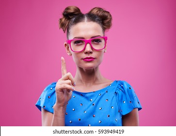 Close-up Of Slim And Good-looking Woman In Pink Glasses Holding Index Finger Up. Female With Two Buns Isolated Over Pink Background 