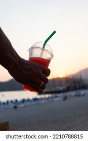 Close-up Of A Slushy In A Plastic Cup With A Green Straw Held By A Man's Hand And In The Background The Sunset On The Beach.