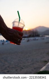 Close-up Of A Slushy In A Plastic Cup With A Green Straw Held By A Man's Hand And In The Background The Sunset On The Beach.