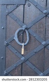 A close-up of a small brass padlock securing a heavy-duty wooden door with decorative black iron cross straps, rivets, and a large metal ring latch.