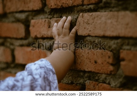 Close-up of a small child’s hand touching a rustic brick wall. The image conveys a sense of exploration, curiosity, and the tactile connection between a baby and their environment.