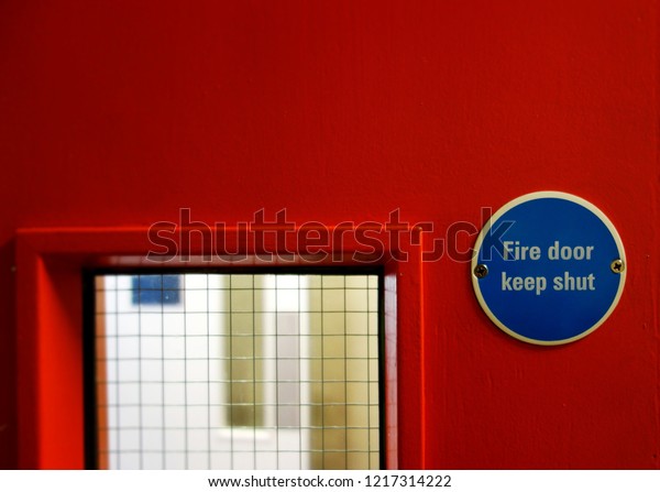 Closeup small warning sign, Fire door keep shut.\
Selective focus on blue metal badge sign on right side of frame.\
Space to add text on red surface, blurry mesh grass & white\
door in backgound. 