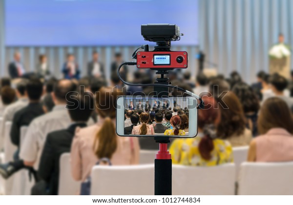 Closeup\
smart mobile phone taking Live over Speakers on the stage with Rear\
view of Audience in the conference hall or seminar meeting,\
technology live streaming and broadcast\
concept