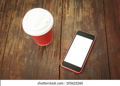 Closeup Of Smart Phone With Isolated White Screen And Takeaway Coffee Cup On Wooden Table Surface. Mockup Concept.