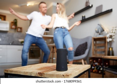 Cierre de un altavoz inteligente sobre una mesa de madera contra una pareja feliz bailando en una sala de estar en casa