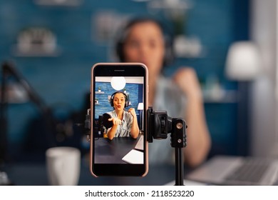 Closeup of smartphone recording woman influencer on live stream podcast. Vlogger talking in front of phone camera and filming video for social media channel, using microphone and headphones.