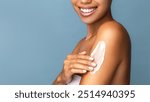 A close-up of a smiling  african woman applying body skin care lotion to her shoulder. The image highlights skincare routine