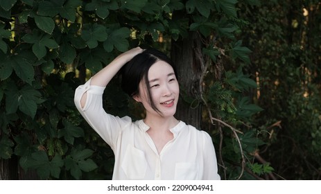 Closeup Of A Smiling Asian Woman Standing Before Green Trees With One Hand Touching Hair. Outdoor Portrait. 