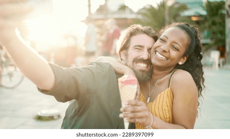 Closeup of smiling interracial couple eating ice cream and taking a selfie on urban city background. Close-up of a man and woman tasting ice cream and video chatting using a mobile phone. Backlight - Powered by Shutterstock