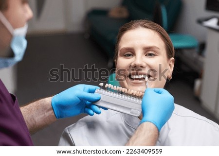 Close-up smiling pretty woman patient in dentist's seat, looking at camera while doctor orthodontist, choosing the shade of dental veneer according to color chart. Aesthetic dentistry. Dental practice
