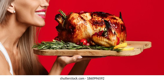 Closeup of smiling sexy blonde woman in holding tasty whole roasted chicken on wooden tray at her face and smells relishes it over red background with copy space. Shape, figure, healthy lifestyle