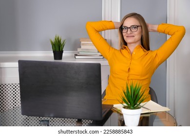 Close-up of a smiling woman in an orange blouse and glasses sitting at a glass table, puts her hands behind her head and looks at a laptop. Concept of success, joy, victory, rest - Powered by Shutterstock