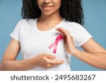 Close-up of smiling young African American woman wearing a pink ribbon pinned to her shirt, symbolizing breast cancer awareness and the importance of early detection. Health care concept