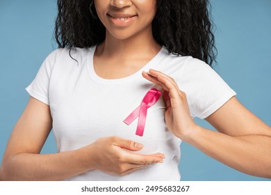 Close-up of smiling young African American woman wearing a pink ribbon pinned to her shirt, symbolizing breast cancer awareness and the importance of early detection. Health care concept - Powered by Shutterstock