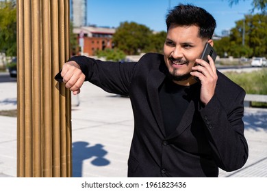 Close-up Of Smiling Young Latino Man Talking On His Cell Phone Or Smart Phone In A Public Square.