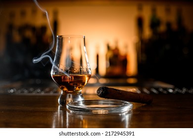 Close-up Of Smoking Cuban Cigar On Ashtray And Glass With Whiskey On Blurred Bar Background