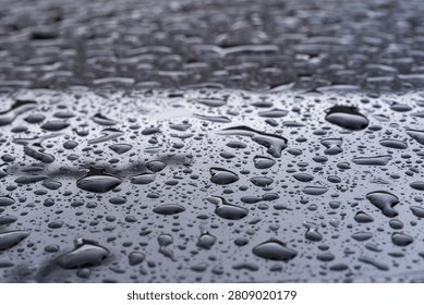 Close-up of a smooth metallic surface covered with water droplets of various sizes. The droplets form an organic pattern, reflecting light and faint shapes from the surroundings, creating a study of surface tension and reflective geometry.