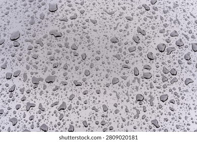 Close-up of a smooth metallic surface covered with water droplets of various sizes. The droplets form an organic pattern, reflecting light and faint shapes from the surroundings, creating a study of surface tension and reflective geometry.