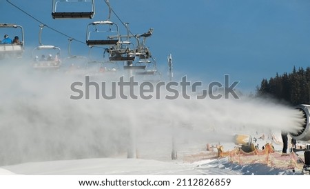 Closeup snow cannons makes snow on sunny day, on bright blue sky. In backdrop skiers use skilift. Snowmaker machine produce artificial snow in Bukovel ski resort. Preparation mountain slope for skiing